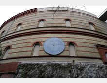 Zylindrisches Backsteingebäude mit roten und gelblichen Streifen, mehreren bogenförmigen Fenstern und einer großen, runden Skala an der Fassade. Im Vordergrund ein Felsvorsprung mit karger Vegetation.