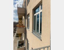 Fassade eines Wohngebäudes mit beigefarbenem Putz, mehreren Fenstern mit weißen Rahmen und mehreren Balkonen mit Geländern. Auf den Balkonen stehen Pflanzkübel und vereinzelt Möbel, im Hintergrund ist ein Gehweg und teilweise bewölkter Himmel zu sehen.