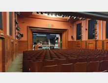 Innenansicht eines Theatersaals mit leeren Reihen aus Holzstühlen, einer Bühne mit Bühnenvorhängen und einer Kulisse im Hintergrund. Die Wände sind in Erdtönen gehalten, mit blauen Akzenten und eingebauten Beleuchtungseinrichtungen.
