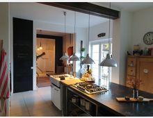 Offene Küche mit Kücheninsel und Hängelampen, modernem Gasherd und dunkler Arbeitsplatte. Im Hintergrund befindet sich ein Durchgang mit einer Schiefertafelwand, hölzernem Dekor und großen Fenstern, die Tageslicht hereinlassen.