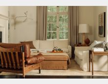 Gemütliches Wohnzimmer mit cremefarbenem Sofa und passender Ottomane, beige Vorhänge vor einem großen Sprossenfenster, brauner Ledersessel im Vordergrund und Holz-Beistelltischen. An der Wand befindet sich eine moderne Wandleuchte und ein abstraktes Kunstbild.