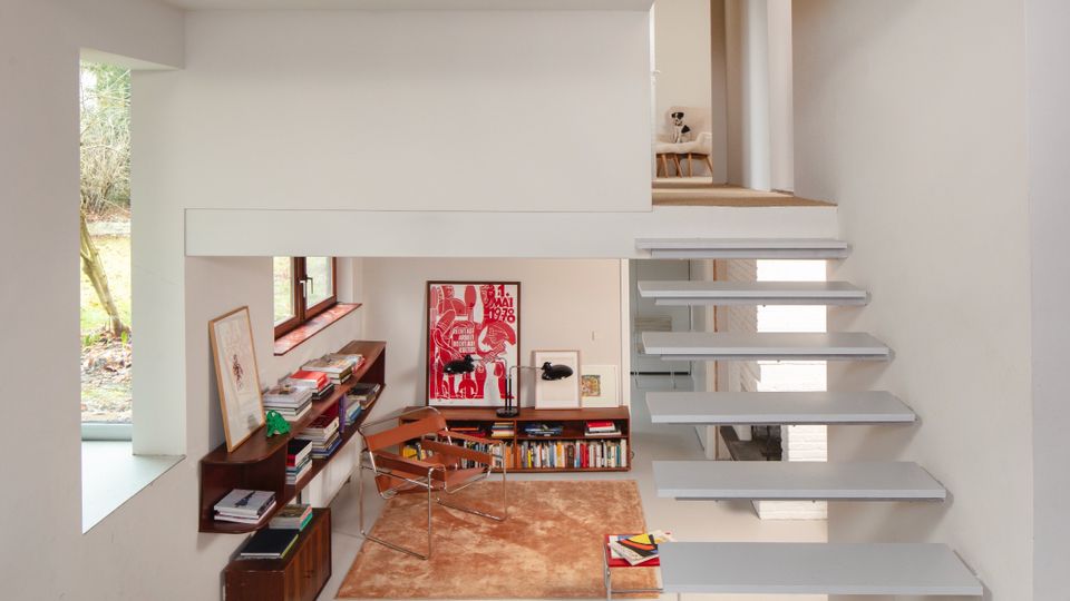 Split-level modern home interior with white walls and a floating gray staircase leading to an upper landing. Below, a small reading or study nook features built-in wood shelving filled with books, framed artwork, and minimalist chairs beside a window.