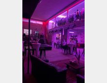 Industriell gestalteter Veranstaltungsraum mit hohen Decken, Ziegelwänden und lila Beleuchtung. Der Raum enthält Sitzgelegenheiten, Stehtische, eine kleine Bühne und eine Empore, auf der Kleidung ausgestellt ist.