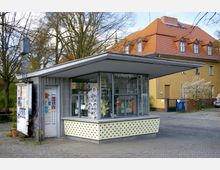Ein kleiner moderner Pavillon mit flachem Dach, großer Glasfront und seitlichen Werbeplakaten, beschriftet mit "KulturKiosk". Im Hintergrund ein gelbes Gebäude mit rotem Ziegeldach und mehrere Bäume.