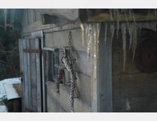 Holzschuppen mit verwitterten Brettern, einem kleinen Fenster und angelehnter Tür. An der Wand hängen ein Seil, eine alte Laterne und Metallgegenstände; von der Überdachung tropfen Eiszapfen.