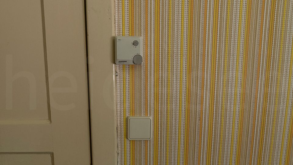 Innenraum mit beige gestrichener Holztür und dunkler Klinke neben einer Wand mit gelb-grau gestreifter Tapete. An der Wand sind ein Lichtschalter und ein kleines Bediengerät montiert; unten ist die Ecke eines gepolsterten Möbels sichtbar.