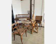Raum mit einer Sammlung alter handwerklicher Werkzeuge und Möbel, darunter ein Spinnrad, ein Webstuhl, eine hölzerne Spule sowie ein antiker Stuhl. Die Gegenstände sind aus Holz gefertigt und stehen vor weißen Wänden auf einem Teppichboden.