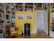 Ein gemütliches Wohnzimmer mit einer gelben Wand, darauf zwei Bilderrahmen. Ein schwarzer Kaminofen steht auf einer Glasplatte, flankiert von einem Korbsessel. Die übrigen Wände sind mit hohen weißen Bücherregalen gefüllt, die bis unter die Decke reichen. Eine Leiter ermöglicht den Zugang zu den oberen Regalböden.