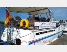 Ein weißes Ausflugsschiff mit gelbem Rettungsring und Korbstühlen auf dem Sonnendeck liegt an einem Steg. An der Seite des Schiffes hängt ein Banner mit der Aufschrift "Feiern · Tagen · Sightseeing" und einer Website-Adresse.