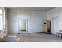 Ein leerer, renovierungsbedürftiger Raum mit abblätternder Wandfarbe, einem braunen Kachelofen in der Ecke, einem Fenster mit Tageslichteinfall auf der linken Seite und zwei Türen; eine führt in einen weiteren Raum mit gefliestem Bereich. Ein Stuhl steht rechts an der Wand.