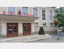 Fassade eines Bibliotheksgebäudes mit heller Putzfassade, großen Fenstern und roter Beschriftung "BIBLIOTHEK" über dem Eingang. Vor dem Eingang befinden sich Stufen, Blumenschmuck und ein gepflasterter Vorplatz.