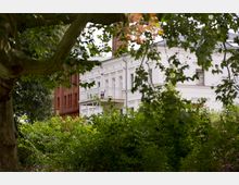 Weißes, historisches Gebäude mit klassischer Fassadengestaltung und Balkon, teilweise verdeckt durch dichte grüne Bäume und Sträucher; rechts daneben ein angrenzendes rotes Backsteinhaus.