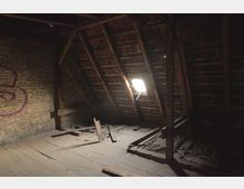 Dunkler, verlassener Dachboden mit freiliegenden Holzbalken und einem kleinen Fenster im Dach, durch das Licht einfällt. Eine Ziegelsteinwand mit Graffiti ist sichtbar, und der Boden ist mit Schmutz und Holzteilen bedeckt.