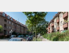 Straßenansicht mit zwei Reihen von mehrstöckigen Wohngebäuden mit roten Ziegeldächern und Balkonen, flankiert von Bäumen und geparkten Autos. Rechts verläuft ein Fußweg mit Grünpflanzen entlang der Häuser.