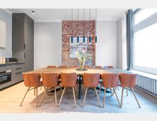 Moderner Essbereich mit großem Holztisch und mehreren braunen Lederstühlen, angrenzend an eine dunkle Einbauküche. Im Hintergrund eine freigelegte Ziegelwand mit Bild, darüber schlanke Pendelleuchten; rechts ein großes Fenster mit Vorhang und Heizkörper darunter.