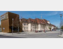 Historisches Gebäude mit heller Fassade und rotem Ziegeldach, daneben ein modernes, würfelförmiges Bauwerk mit goldener Metallverkleidung. Im Vordergrund befinden sich Bäume, Fahrradständer und Laternen, im Hintergrund ein hoher Sendemast.