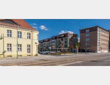 Straßenansicht mit einem historischen Gebäude links mit gelber Fassade und rotem Dach, modernen Wohnhäusern mit Balkonen im Hintergrund und einer Straßenbahnhaltestelle sowie Oberleitungen im Vordergrund. Der Himmel ist klar und blau.