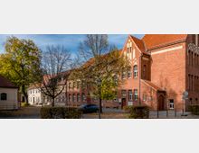 Ziegelsteinbau mit roten Dachziegeln und gotischen Stilelementen in einem urbanen Umfeld, umgeben von Bäumen mit herbstlich gefärbtem Laub; im Vordergrund eine kleine Grünfläche und ein parkendes Auto.