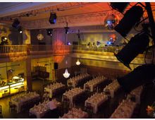 Innenraum eines Veranstaltungssaals mit festlich gedeckten Tischen in mehreren Reihen. Oben befindet sich eine Galerie mit Geländer, dekoriert mit Kronleuchtern und Discokugeln; Scheinwerfer und Bühnentechnik sind an der Decke montiert.