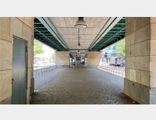 Unterführung eines Hochbahnviadukts mit gemauerten Pfeilern und einer grünen Stahlkonstruktion an der Decke. Auf beiden Seiten sind Fahrradständer und im Hintergrund sind Stadtgebäude sowie vorbeifahrende Fahrzeuge zu sehen.