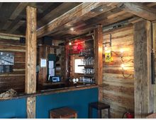 Holzvertäfelte Bar in rustikalem Stil, mit sichtbaren Holzbalken, einer Theke mit Gläsern und Schankhähnen sowie Wandleuchten im Vintage-Design. Auf der Theke steht ein Korb mit Brot, und die Umgebung wirkt gemütlich und traditionell.