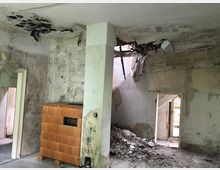 Ein verfallener Innenraum mit beschädigten Wänden und Decken, sichtbarem Schutt am Boden und einem freistehenden, braunen Kachelofen. Die Decke weist ein großes Loch mit herabhängenden Trümmern auf, und die Wände sind von Schimmel und Feuchtigkeit gezeichnet.