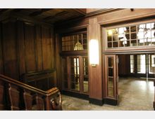 Holzvertäfeltes Treppenhaus mit Glastüren und einem Fenster im oberen Bereich. Der Boden besteht aus Steinfliesen, und an der Wand hängt eine moderne, vertikale Wandlampe.