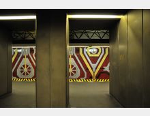 Unterirdischer Durchgang mit metallischen Wänden und grafisch gestalteter Wand am Ende, die rote, gelbe und weiße geometrische Muster sowie technische Motive zeigt. Die Beleuchtung ist künstlich, mit gelblichen Lichtleisten an der Decke.