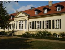 Ein hellgelbes Herrenhaus mit rotem Satteldach, Gaubenfenstern und weißen Fensterrahmen mit dunklen Fensterläden. Die zentrale Eingangspartie verfügt über Säulen und einen Dreiecksgiebel, davor befindet sich ein kleiner Treppenaufgang. Im Vordergrund ist ein Garten mit Rasen und Sträuchern zu sehen.