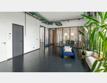 Moderner Büroraum mit dunklem Bodenbelag, weißen Wänden und sichtbaren Kabeltrassen an der Decke. Im Vordergrund ein blauer Sessel sowie Pflanzen, im Hintergrund verglaste Konferenzräume mit runden Hängelampen und Vorhängen.