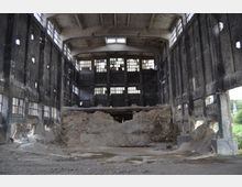 Industriehalle im Verfall mit hohen, dunklen, beschädigten Wänden und großen, vergitterten Fenstern. Der Boden besteht aus Schutt und Erde, während der hintere Bereich stark eingestürzt ist, mit sichtbarem Beton und offenem Himmel durch Löcher in der Struktur.