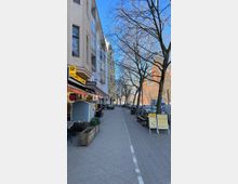 Straßenszene in einer Stadt mit mehrstöckigen Wohnhäusern auf der linken Seite, darunter kleine Geschäfte und Cafés mit Außensitzplätzen. Rechts stehen Bäume ohne Laub, Fahrräder und parkende Autos entlang eines Bürgersteigs.