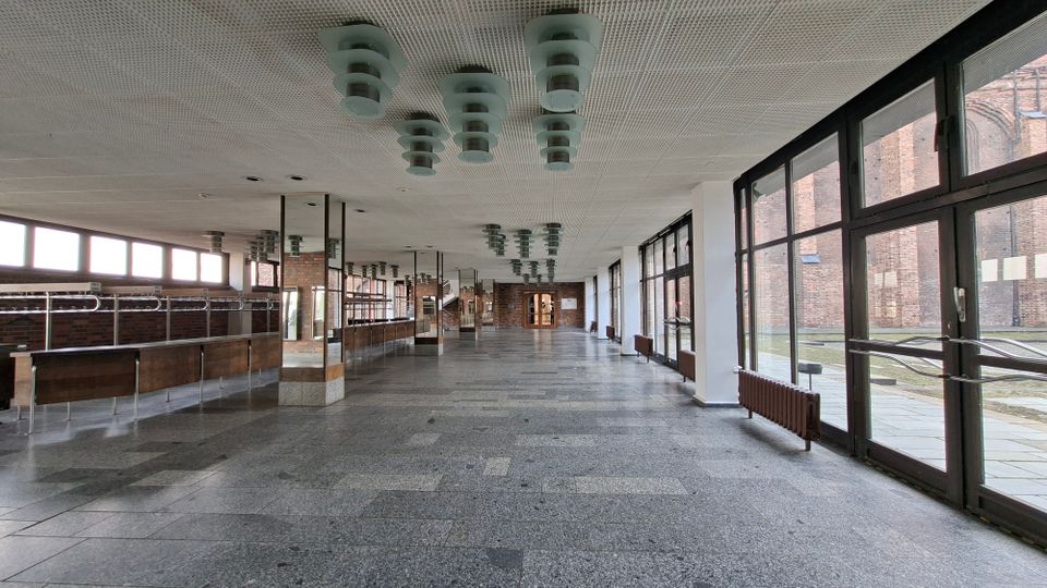 Langer, leerer Innenflur mit grauem Steinboden, weißer Rasterdecke und mehreren zylindrischen Deckenleuchten sowie Spiegel- und Stützsäulen entlang der Mitte. Rechts verläuft eine durchgehende Glasfront mit Türen und Heizkörpern, links stehen lange Theken, im Hintergrund sind Backsteinwände und ein Ausgang zu sehen.