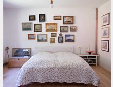 Schlafzimmer mit schlichtem Holzfußboden und weißer Wand, dekoriert mit mehreren maritimen Gemälden in verschiedenen Rahmen. Ein Doppelbett mit geblümtem Bettüberwurf steht zentral, daneben ein Nachttisch mit Lampe und einem Modellschiff.