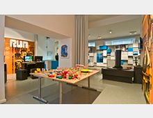 Ein moderner Museumsraum mit interaktiven Ausstellungsstücken, darunter ein Tisch mit Holzbausteinen und ein Computerarbeitsplatz. Im Hintergrund sind farbige Wandtafeln, Grafiken und Vitrinen mit Exponaten zu sehen.