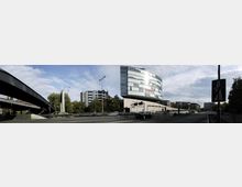 Moderne Bürogebäude mit Glasfassade an einer mehrspurigen Straße, daneben Wohnhäuser und Bäume; im Vordergrund eine Brücke aus Stahl, die über die Straße führt. Der Himmel ist teilweise bewölkt, und es sind Verkehrszeichen sowie Autos auf der Straße zu sehen.