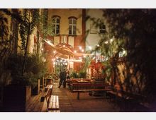Beleuchteter Hinterhof mit Holzterrasse, umgeben von mehreren Sitzbänken und Pflanzen in Töpfen. Lichterketten sind über den Hof gespannt, und im Hintergrund ist ein Gebäude aus Backstein mit hohen Fenstern zu sehen.