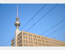 Der Berliner Fernsehturm mit seiner markanten Kugel und der rot-weißen Antenne ragt über einem beige-farbenen Gebäude mit zahlreichen rechteckigen Fenstern, vor einem klar blauen Himmel. Stromleitungen verlaufen diagonal durch das Bild.