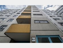 Hochhausfassade mit hellbraunen Kacheln und blauen Fenstern, aus der Froschperspektive aufgenommen. Auffällig sind gelb gestrichene Balkone mit Maschendraht und eine Eingangstür mit der Hausnummer „40 G“.