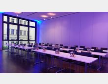 Ein moderner Seminarraum mit weißen Tischen und schlichten schwarzen Stühlen, die in Reihen angeordnet sind. Der Raum ist durch große Fenster mit Blick auf ein gegenüberliegendes Gebäude erhellt, während die Wandbeleuchtung in violettem Licht gestaltet ist.