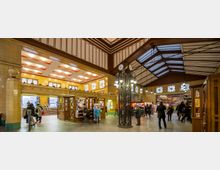 Historischer Bahnhofseingang mit gelb gefliesten Wänden, einer hohen Holz- und Glasdecke, und einer zentralen Uhrensäule. Menschen bewegen sich durch den Raum, in dem sich Informationsstände, ein Kiosk und Zugangsschilder zu den Gleisen befinden.