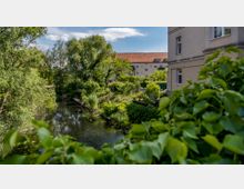 Blick auf einen kleinen, von Bäumen und Sträuchern umgebenen Wasserkanal in einer Wohngegend mit einem beigefarbenen Gebäude und roten Ziegeldächern im Hintergrund. Im Vordergrund sind grüne Pflanzen und Blätter zu sehen.