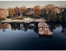 Wide view of a calm river with a large floating wooden deck and dock in the foreground, reflecting trees and nearby buildings in the water. On the opposite bank, terraced walkways and stairs run through autumn-colored trees alongside residential buildings.