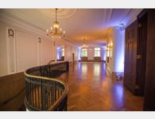 Großer, eleganter Saal mit Parkettboden, verzierten weißen Wänden und kunstvollen Kronleuchtern an der Decke. Ein geschwungenes Geländer führt zu einer Treppe, während Fenster an der Stirnseite des Raumes natürliches Licht einlassen.