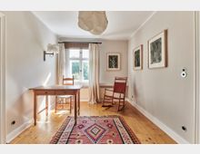 Helles Zimmer mit Holzboden, einem kleinen Holztisch und zwei Stühlen (einem einfachen und einem Schaukelstuhl). An der Wand hängen drei gerahmte Bilder, vor dem Tisch liegt ein gemusterter Teppich, und ein Fenster mit Gardinen lässt Tageslicht herein.