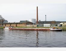 Industriegebiet mit einer Wasserstraße im Vordergrund, auf der ein langes Frachtschiff mit geschlossenen Laderäumen fährt. Im Hintergrund befinden sich Ziegelgebäude, moderne Industriebauten, ein hoher Schornstein und ein weißer zylindrischer Tank.