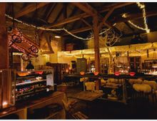 Gemütlicher Innenraum eines rustikalen Lokals mit Holzbalken, freiliegenden Backsteinwänden und zahlreichen warmweißen Lichterketten. Der Raum ist dekoriert mit antiken Gegenständen, hölzernen Möbeln, Schaffell bedeckten Hockern sowie kleinen roten Kerzenleuchten auf den Tischen.