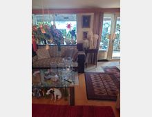 Wohnzimmer mit gemütlicher Einrichtung, bestehend aus einem Sofa mit gestreiften Kissen, einem Glastisch mit dekorativen Objekten und einer Weihnachtsdekoration mit Tannenzweigen. Im Hintergrund sind große Fenster und Glastüren zu einem Außenbereich mit Pflanzen sichtbar, daneben hängen Bilder und Vorhänge an den Wänden.