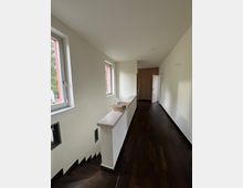 Ein schmaler, heller Flur mit dunklem Holzfußboden, weißen Wänden und drei seitlich angeordneten Fenstern. Links unten führt eine Treppe nach unten, und am Ende des Flures befindet sich eine geschlossene Tür aus Holz.