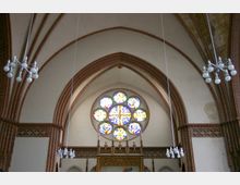 Innenansicht einer Kirche mit gotischen Spitzbögen aus roten Ziegeln, einem verzierten Altar und einem großen runden Fenster mit bunten Glasmalereien. Von der Decke hängen moderne Lampen mit kugelförmigen Leuchten.